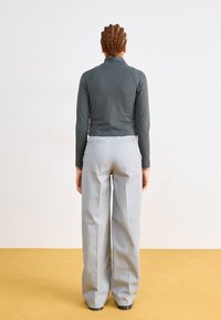 Haut gris à manches longues avec col haut, associé à un pantalon large gris clair, présentant des poches arrière et un léger éclat, sur un sol jaune.