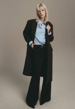 Abrigo negro oversized sobre una camisa de rayas azul claro y blanca, combinado con pantalones negros de tiro alto y zapatos negros de punta.