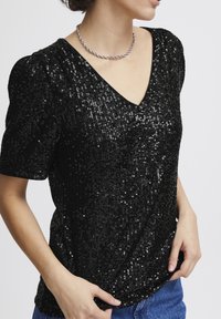 Svart paljett-topp med V-ringning och korta puffärmar. Mjuk textur, glänsande yta med en figursydd design, bärs över blå jeans.