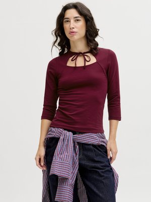Ženska z valovitimi temnimi lasmi, oblečena v bordo majico z dolgimi rokavi s sprednjim vozlom, temne hlače in črtasto srajco, vezano okoli pasu.