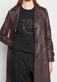 Cappotto in pelle color bordeaux sopra una felpa nera con applicazioni di strass. Pantaloni neri completano il look.