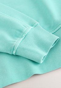 Détail de la manche à revers d'un sweat-shirt turquoise clair montrant un poignet et un ourlet en maille côtelée avec couture visible.