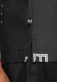 T-shirt de sport noir avec une fente latérale, une étiquette de marque blanche, et un motif géométrique blanc sur fond noir. Confectionné en matière légère.