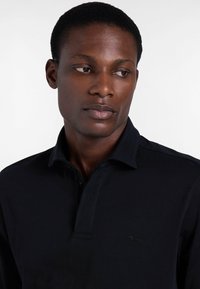 Schwarzes Polohemd mit Kragen, ausgestattet mit einer vorderen Knopfleiste und einem kleinen gestickten Logo auf der Brust. Glatte Stoffstruktur.