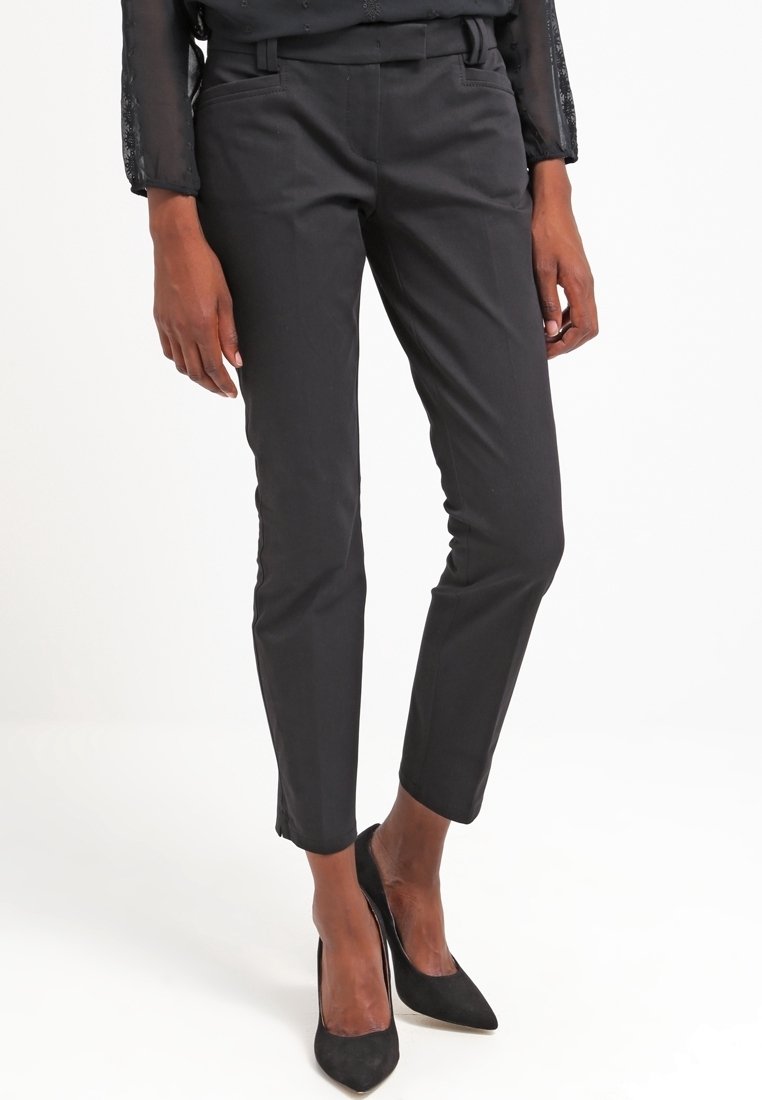 Pantalon noir sur mesure avec une coupe slim, doté d'un devant plat, de passants pour ceinture et d'une texture lisse. Porté avec des chaussures noires à talons hauts.