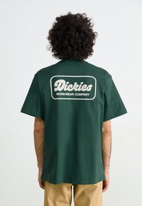 Grøn kortærmet T-shirt lavet af bomuld med en afslappet pasform. Har et stort hvidt logo på ryggen med teksten "Dickies Workwear Company."