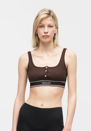 Blonde vrouw die een bruin cropped tanktop met knopen en "Versace" logo-band draagt, gecombineerd met zwarte high-waisted broek, neutrale achtergrond.