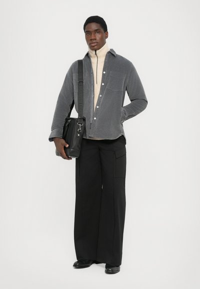 Mann in grauem Cordjackett über beiger Rollkragenpullover, schwarzen weitem Hosen, schwarzen Schuhen und mit schwarzer Umhängetasche.
