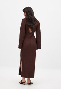 Robe brune texturée avec des manches longues, un dos ouvert et des fentes sur les côtés. Le tissu a un motif froissé, offrant une silhouette ajustée.