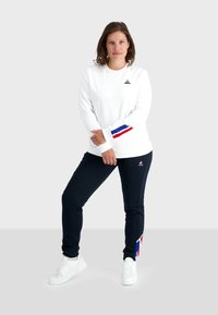 Felpa bianca con accenti a righe rosse e blu sulle maniche, abbinata a joggers blu scuro con accenti simili e un dettaglio con logo.