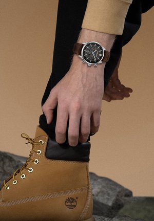 Orologio con cinturino in pelle marrone, quadrante nero e dettagli argento, indossato su una mano accanto a un stivale beige con colletto nero e occhielli dorati.