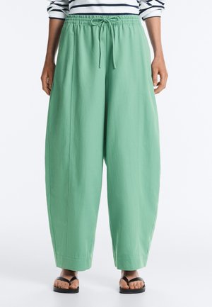 Person trägt lockere, hoch taillierte mintgrüne Hose mit Kordelzug, kombiniert mit schwarzen Flip-Flops, sichtbares gestreiftes Cropped-Top.