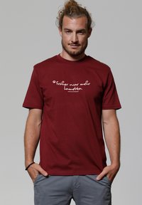 Man met een baard en vastgebonden haar, gekleed in een rode t-shirt met de Duitse tekst "früher war mehr lametta" en grijze broek, met zijn handen in zijn zakken.