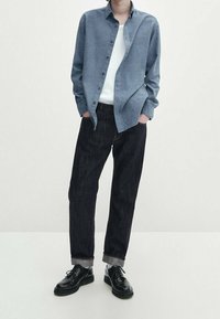 Helles blaues Button-Down-Hemd, weißes Unterhemd, dunkle Jeans und schwarze Schnürschuhe. Glatter Stoff, entspannte Passform und umgekrempelte Säume.