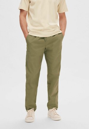 Pantaloni leggeri verde oliva con vita elastica, tasche laterali e vestibilità comoda, abbinati a una t-shirt beige e scarpe da ginnastica chiare.