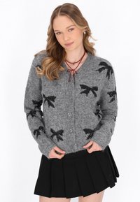 Cardigan grigio a maglia con motivi a fiocco neri, cinque bottoni e maniche lunghe. Abbinato a una gonna nera plissettata.