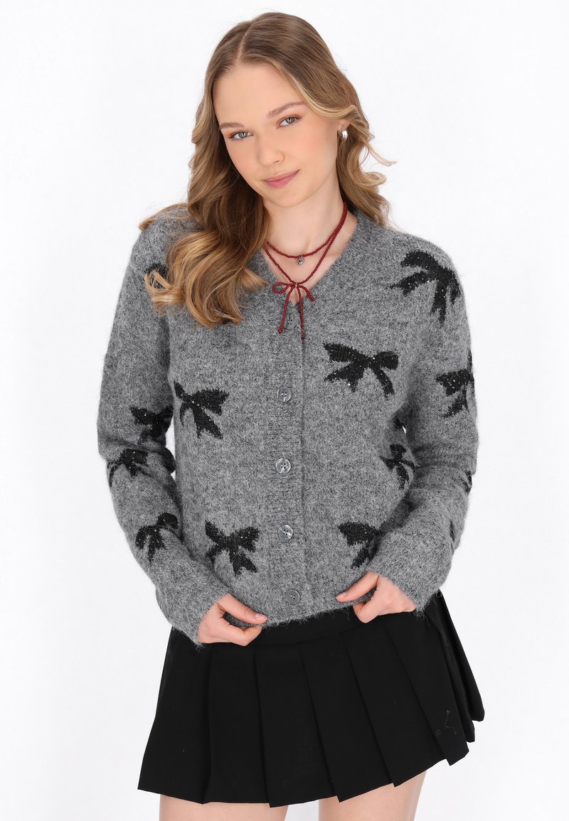 Cardigan grigio a maglia con motivi a fiocco neri, cinque bottoni e maniche lunghe. Abbinato a una gonna nera plissettata.