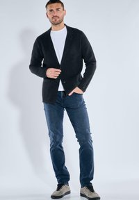 Mann, der einen schwarzen Cardigan, ein weißes T-Shirt, eine blaue Jeans und graue Turnschuhe trägt und vor einem schlichten hellgrauen Hintergrund steht.