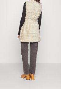 Beżowa kamizelka z sztucznego futra z paskiem w talii, z teksturowanym paskowanym wzorem, połączona z ciemnoszarymi szerokimi spodniami dżinsowymi i pomarańczowymi butami na słupku.