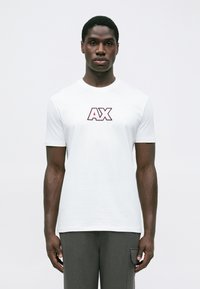 Mladic u bijelom majici s velikim "AX" logotipom i sivoj hlačama stoji ispred jedne obične bijele pozadine.