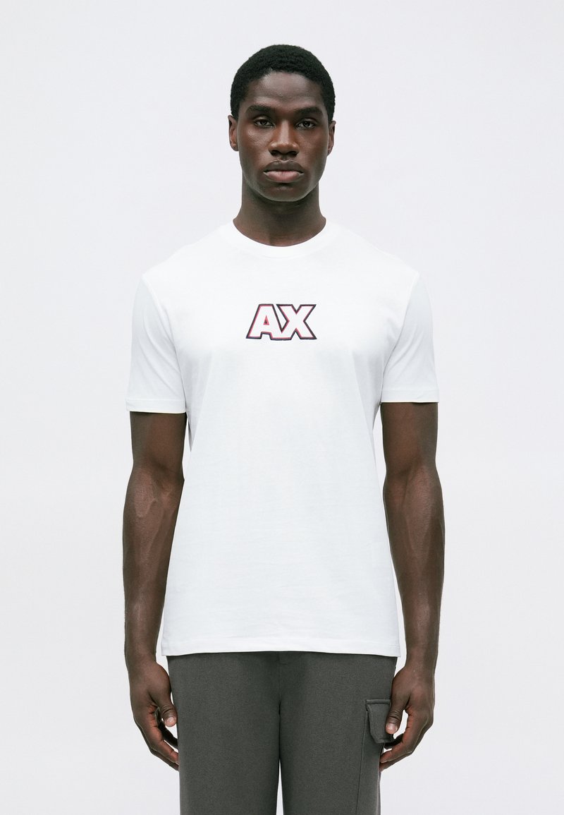 Mladic u bijelom majici s velikim "AX" logotipom i sivoj hlačama stoji ispred jedne obične bijele pozadine.
