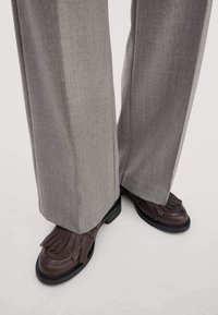 Bruine leren schoenen met franjes, zwarte zool, gedragen met grijze wijde broek, met een gladde textuur en zorgvuldig ontworpen pasvorm.