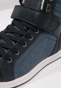 Vysoké tenisky s designem z tmavě modrých smíšených materiálů, obsahující strukturované kožené tělo, černé tkaničky a suchý zip pro zapínání.
