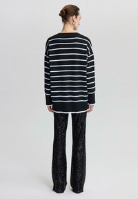 Kvinna som står vänd bort, iklädd en lång tröja med svartvita horisontella ränder och svarta paljettbyxor, håret i en knut.