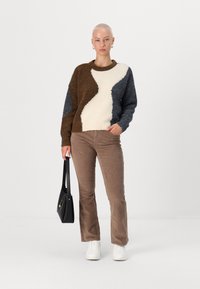 Pull en tricot marron, crème et bleu marine ; présente un design abstrait. Associé à un pantalon en velours côtelé beige et des baskets blanches. Porte un sac à main noir.