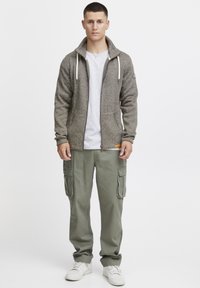 Grå strikket zip-up jakke med snoretræk, parret med løstsiddende grønne cargobukser med sidelommer. Båret med hvide sneakers.