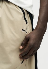 Σκούρο δέρμα χέρι που ακουμπά κοντά στην τσέπη σορτς Puma μπεζ με μαύρο φερμουάρ και λογότυπο Puma, φορώντας άσπρο πουκάμισο.