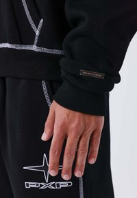 Sudadera negra con detalles de costura blanca. Presenta puños acanalados y una pequeña etiqueta metálica con el logo que dice "PROJECT PARENT."