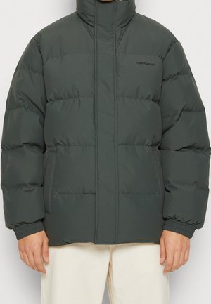 Dunkelgrüne, wattierte Winterjacke mit hohem Kragen, elastischen Bündchen, Fronttaschen und dezentem Carhartt-Logo auf der Brust, getragen zu hellen Hosen.