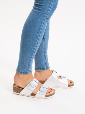 Feet wearing white slip-on sandals with blue buckles and cork soles paired with blue skinny jeans.