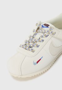 Hvit Nike-sneaker med fargerike mønstrede lisser, beige såle og en liten flerfarget brodert Nike-swoosh på tåområdet.