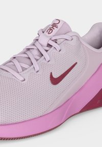 Ανοιχτό μοβ αθλητικό παπούτσι Nike με ροζ σόλα, κόκκινο λογότυπο Swoosh και πάνω μέρος από δίχτυ με δεμένα κορδόνια σε λευκό φόντο.
