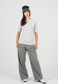 Donna che indossa un cappellino nero, una maglietta grigia chiara a maniche corte, pantaloni grigi a gamba larga e mocassini bianchi, in piedi contro uno sfondo semplice.