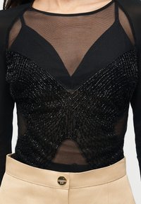 Μαύρο διάφανο τοπ με διακοσμητική πεταλούδα από χάντρες πάνω από μαύρο σουτιέν, συνδυασμένο με μπεζ ψηλόμεσο παντελόνι με χρυσό κούμπωμα.