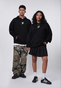 Felpe nere con accenti grafici bianchi, abbinate a pantaloni con motivo mimetico e una gonna nera plissettata. Sneakers nere completano il look.