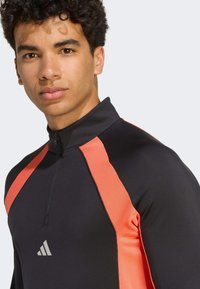 Athletic long-sleeve top in black with coral accent stripes, featuring a high collar, half-zip closure, and textured fabric details.