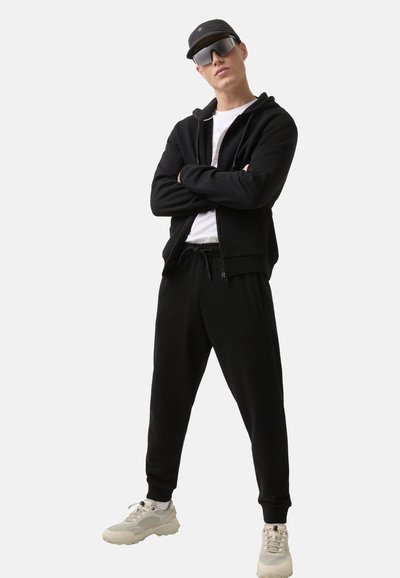 Schwarzer Reißverschluss-Hoodie über einem weißen T-Shirt, kombiniert mit schwarzen Jogginghosen und hellgrauen Turnschuhen. Accessoires sind eine schwarze Kappe und Sonnenbrille.