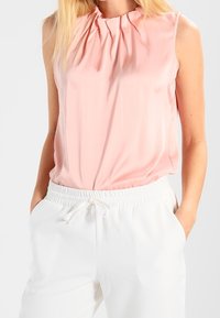 Blouse rose sans manches avec encolure plissée, fabriquée dans un tissu lisse. Associée à un pantalon blanc doté d'une taille élastique et d'un cordon de serrage.