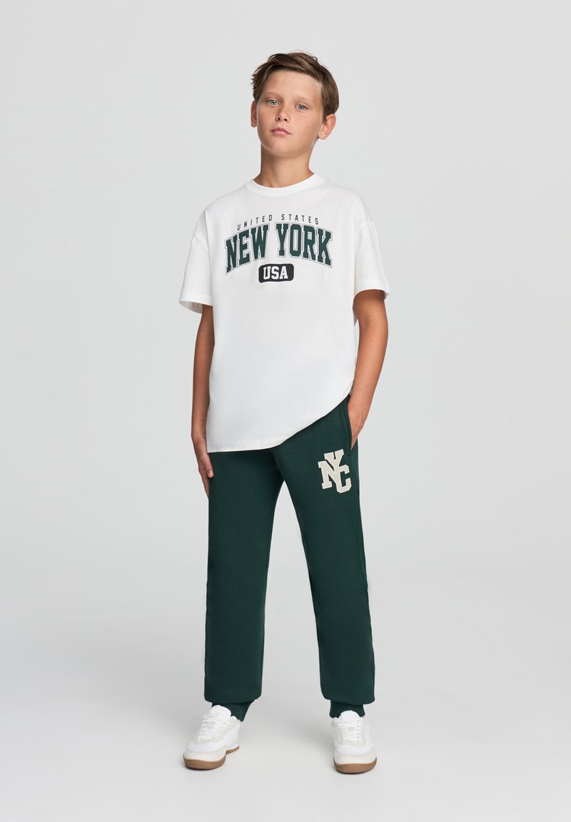 Biała koszulka z napisem "NEW YORK" w kolorze zielonym, połączona z ciemnozielonymi spodniami dresowymi z beżowym logo "NY". Całość dopełniają białe sneakersy.