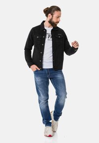 Veste en denim noir avec poches avant, portée par-dessus un t-shirt blanc. Associée à un jean bleu et des baskets blanches avec des accents rouges.