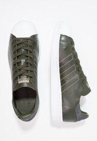 Tmavě zelené kožené tenisky s bílým gumovým špičkou a podešví, s béžovými bočními pruhy a odpovídajícími tkaničkami. Zahrnuje detail ze semišové kůže na patě.