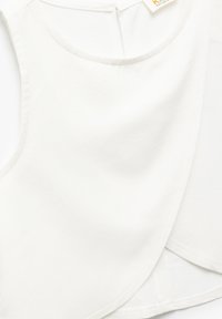 Haut sans manches blanc, en tissu doux avec un col rond. Présente un design croisé à l'avant et une fermeture en trou de serrure au dos.