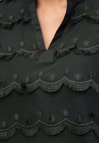 Tecido preto com franjas em bordado em escamas e padrões circulares, apresentando um decote em V na parte superior do peito de uma pessoa.