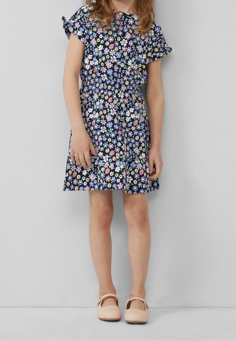 Jeune fille portant une robe florale bleu marine à manches courtes et des chaussures Mary Jane roses, debout devant un fond gris uni.