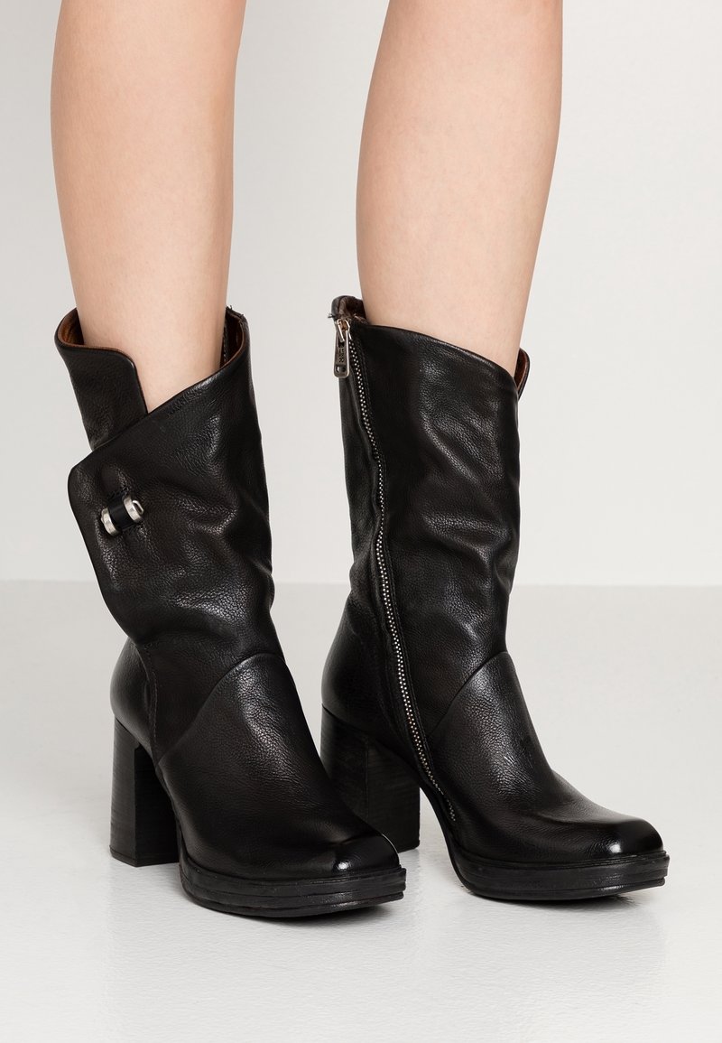 Svarta läderstövlar i midiklass med blockklackar, en stövel har en synlig sidogädda och den andra är vikt över med ett dekorativt spänne.