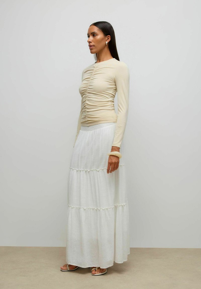 Moteris su ilgais tamsiais plaukais, dėvinti smėlio spalvos susmulkintą ilgomis rankovėmis palaidinę, baltą sluoksniuotą ilga sijoną, smėlio spalvos sandalus, ovalius auskarus ir masyvų apyrankę, stovinti šonu.